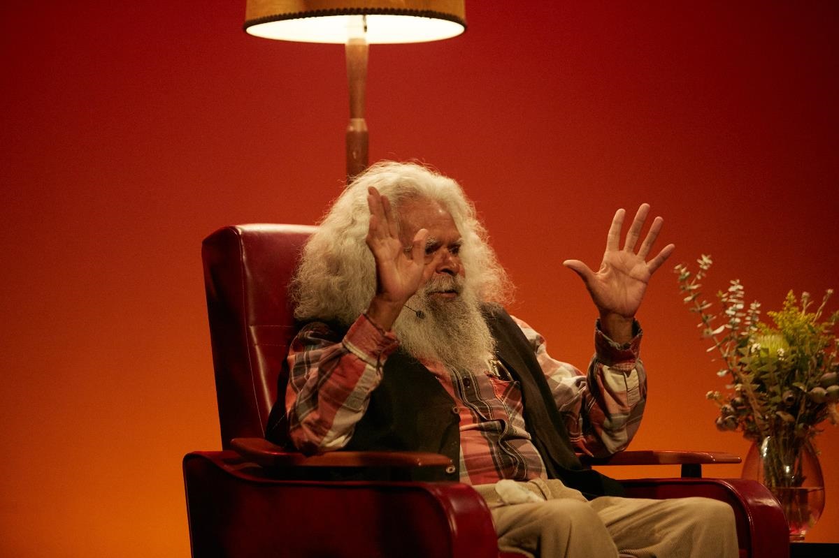 Uncle Jack Charles sits in an armchair with a lamp behind him, as he speaks to the audience.