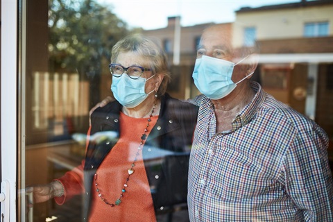 Two older adults look through a window, while wearing face masks.