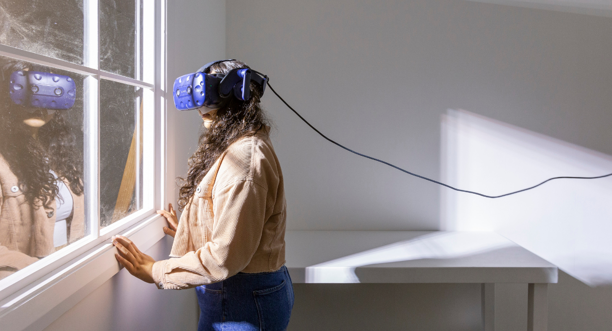 A person with long, curly hair stands in an empty room, looking out of a window. They are wearing a virtual reality headset.