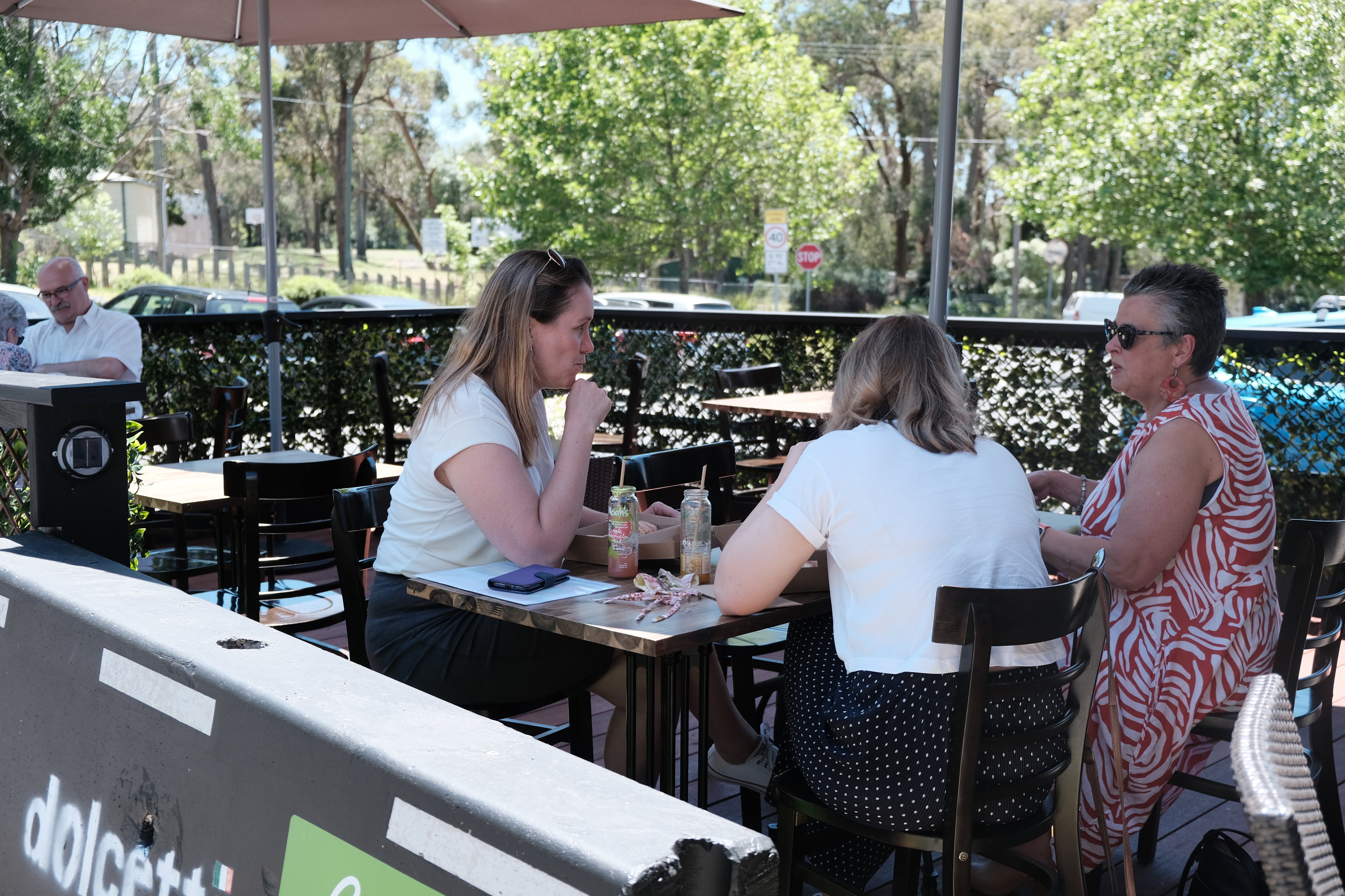 An image of three women in an outdoor dining area, drinking coffee with greenery in the background.