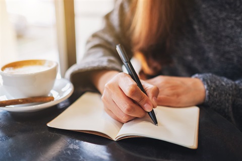 Women writing in a notebook at a table with a cup of coffee next to her