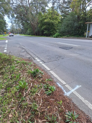 Cambridge Road, Mooroolbark - Roadside view of a two-lane suburban road bordered by trees and vegetation. Cracked and patched asphalt is visible along the road edge, with a marked area on the shoulder in the foreground. A bus stop shelter is visible on the right and a vehicle is travelling away in the distance.
