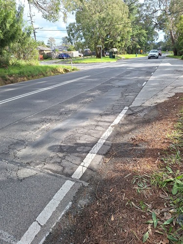 Cambridge Road, Mooroolbark. Two-lane suburban road lined with mature trees and residential properties. Extensive cracking and patching are visible along the road shoulder and traffic lane. A bus stop shelter, roadside drainage channel, and several vehicles are visible further along the road.