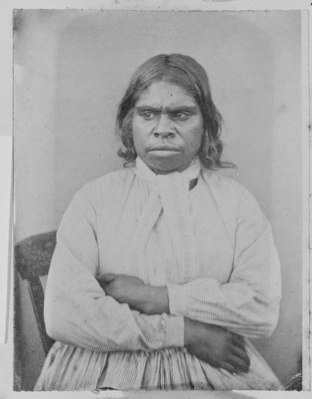 A black and white photograph of a First Nations woman sitting on a wooden chair.
